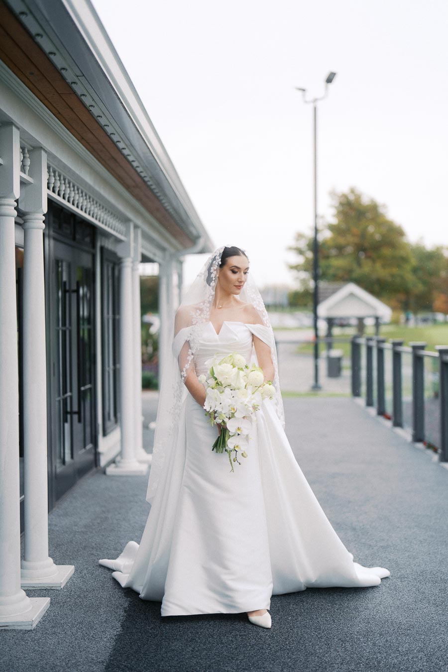 Elegant bride in a white wedding dress holding a bouquet of white flowers, standing outdoors near a classic building with pillars and a glass facade.