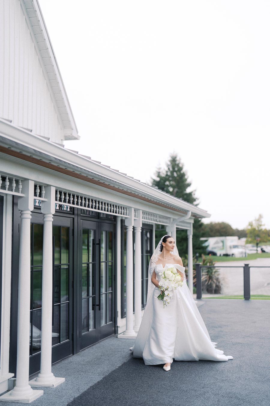 Elegant bride in a white wedding dress holding a bouquet of white flowers, standing next to a modern building with glass doors and columns, surrounded by a landscaped outdoor setting.
