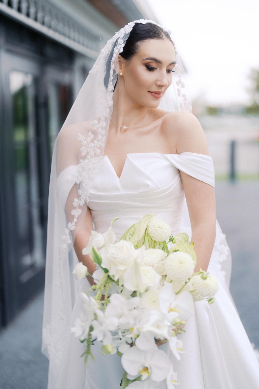 Bride in an elegant off-shoulder white wedding gown with lace veil, holding a bouquet of white flowers, looking down with serene expression.