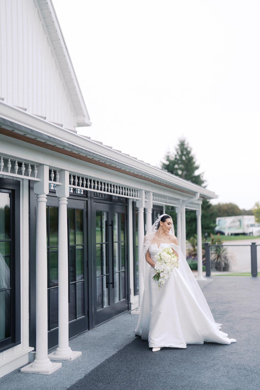 Bride in an elegant white gown holding a bouquet of white flowers, standing outside a modern venue with white pillars and black-framed glass doors.