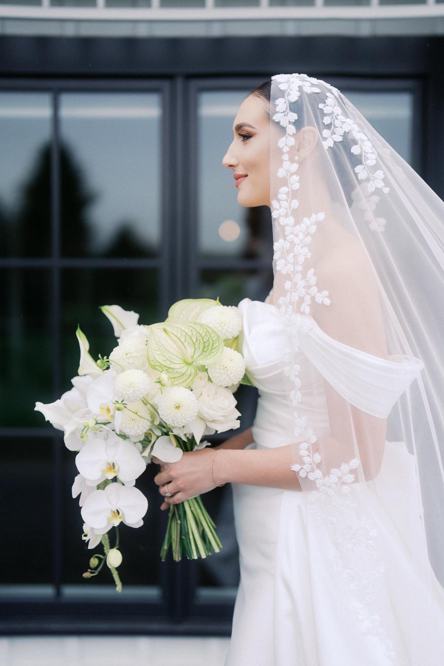 Bride in elegant white gown and floral lace veil holding a bouquet of white flowers and greenery in front of a glass window.