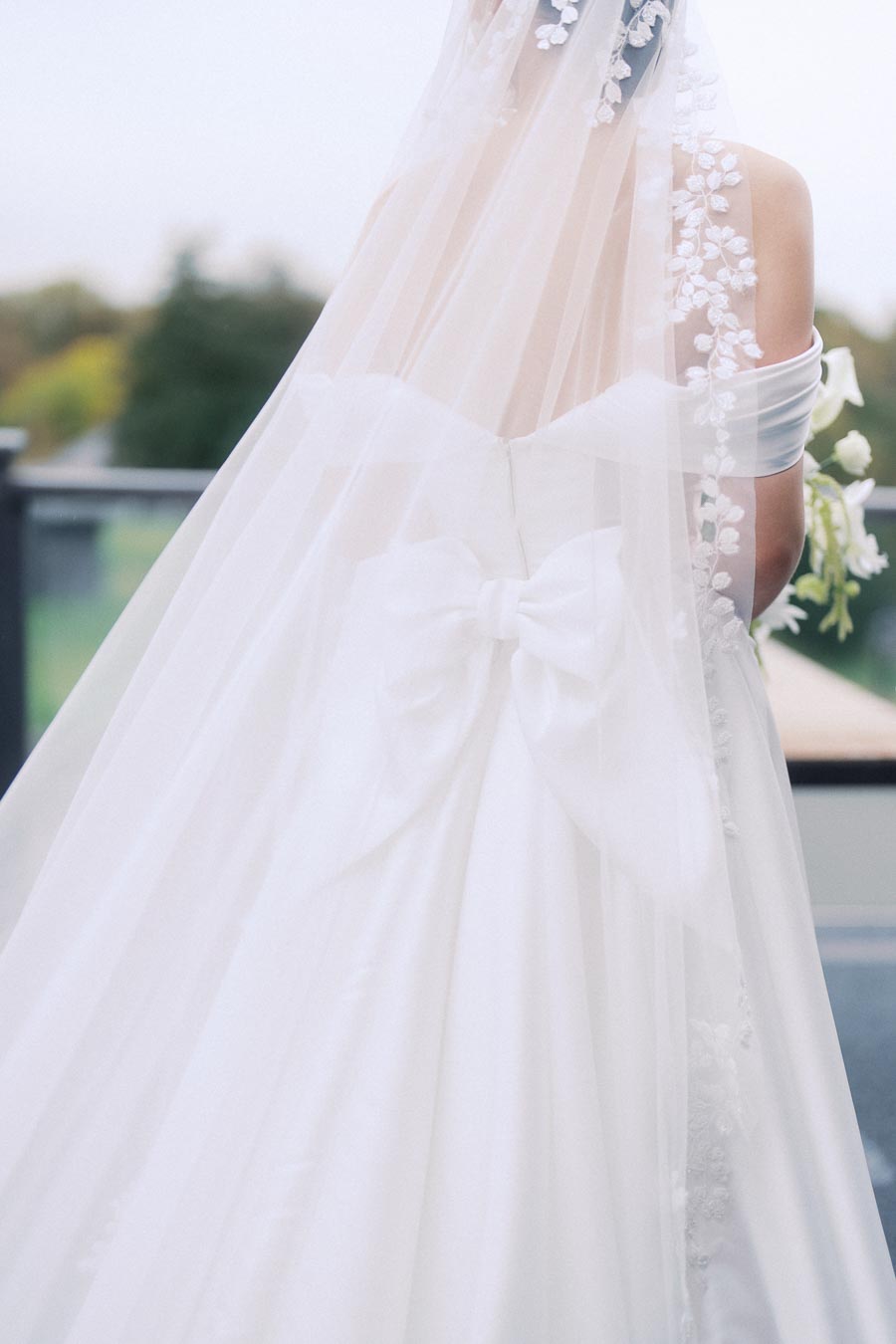 Elegant bridal gown with floral lace veil and large bow on the back, ideal for a romantic outdoor wedding.
