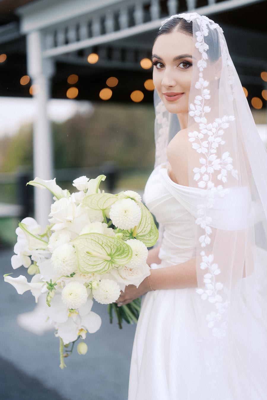 Elegant bride in a white gown holding a bouquet of white and green flowers, wearing a floral lace veil, poses outdoors on a wedding day.