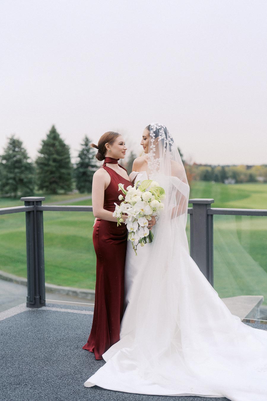 Bride in a white dress with veil holds a bouquet of white flowers while standing next to a woman in a red dress, with a green outdoor landscape in the background.