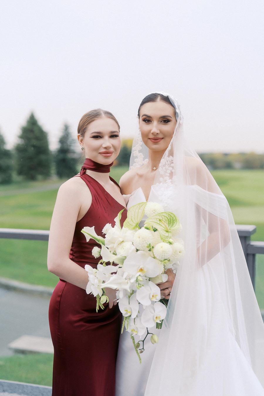 Bride and bridesmaid posing together outdoors, with the bride in a white gown and veil holding a bouquet of white flowers.
