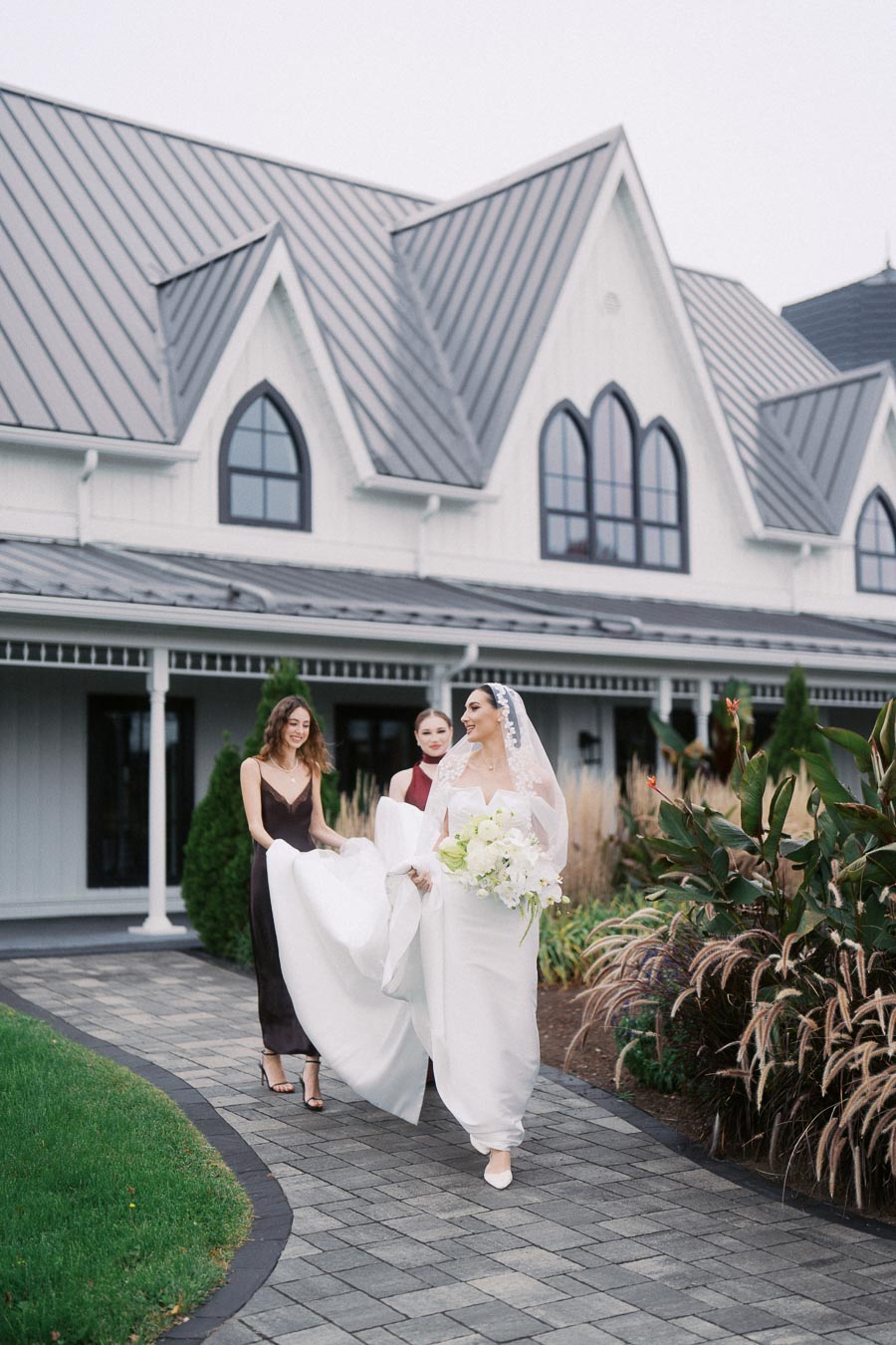 Bride walking with bridesmaids in elegant dresses, carrying a bouquet of white flowers, outside a wedding venue with pointed arched windows and a lush garden backdrop.