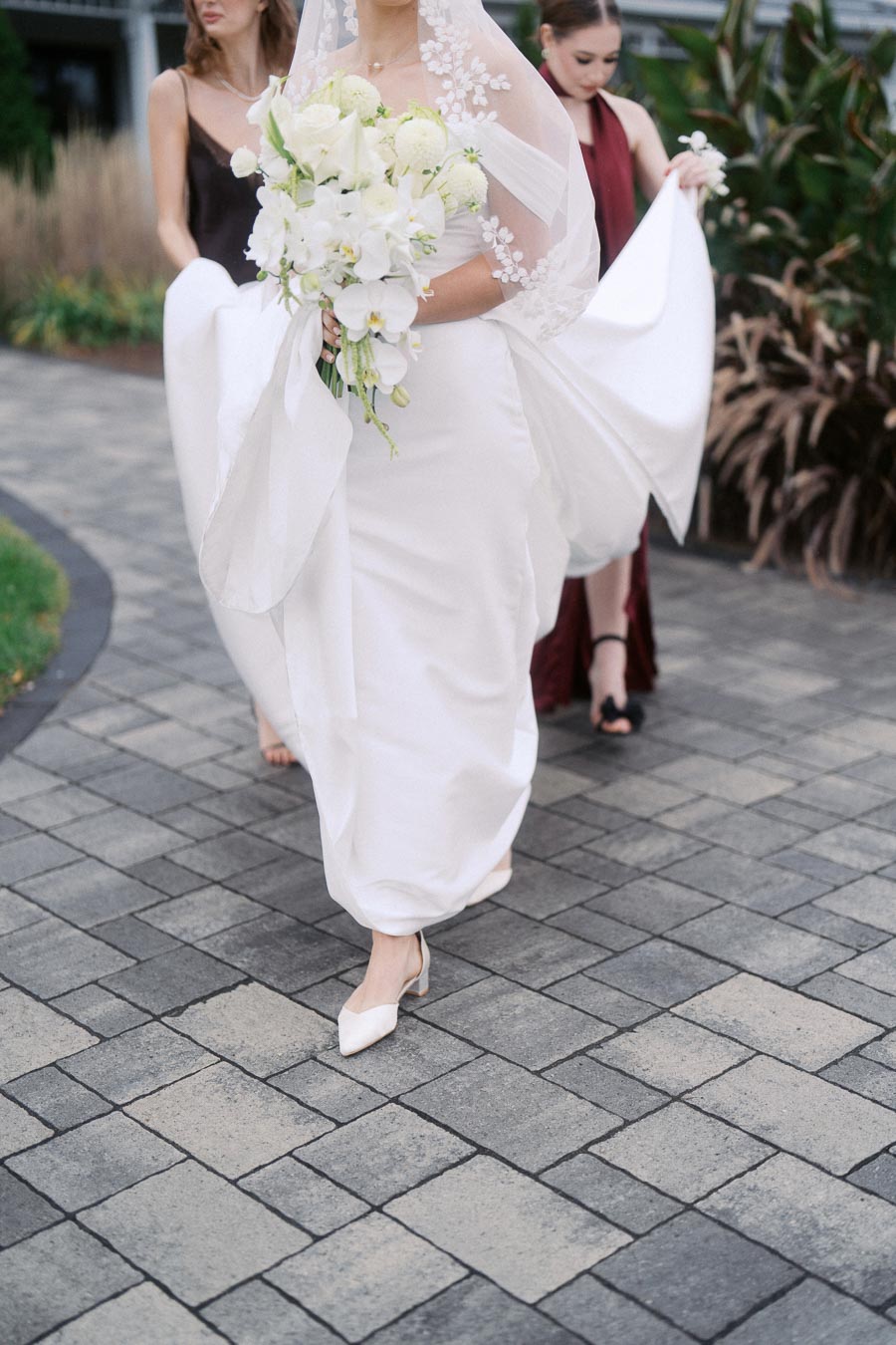 Bride in elegant white gown holding a bouquet of white flowers, walking on a stone pathway with bridesmaids assisting her dress.