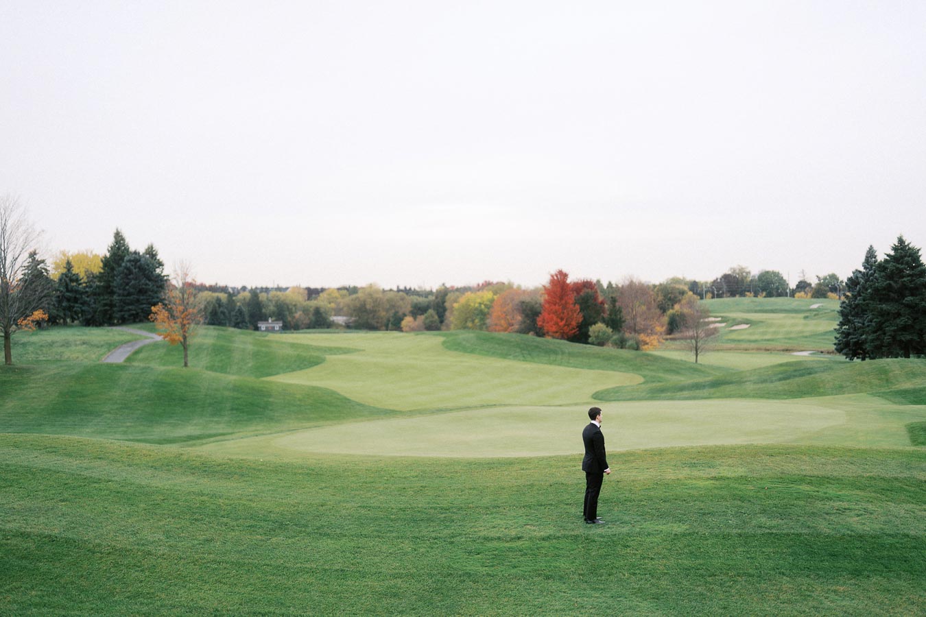 Man in suit standing on a vast, green golf course with rolling hills and colorful autumn trees in the background.