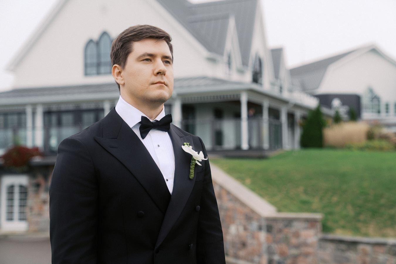 A groom in a black tuxedo with a white boutonniere stands confidently in front of an elegant venue with a lush green lawn, exuding sophistication and style on his wedding day.