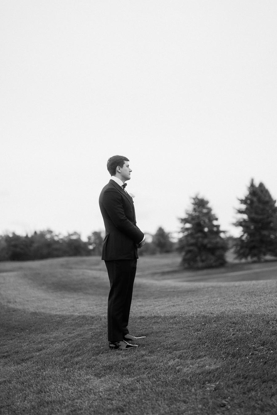 A groom in a black suit stands thoughtfully on a grassy hill with trees in the background, captured in a serene black-and-white photograph.