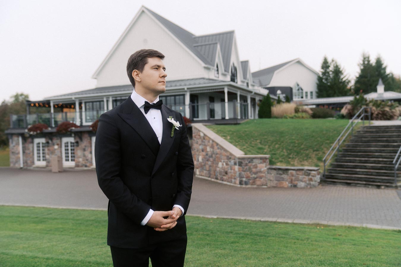 A man in a black tuxedo stands confidently in front of an elegant venue with a landscaped garden, perfect for a formal gathering or wedding event.