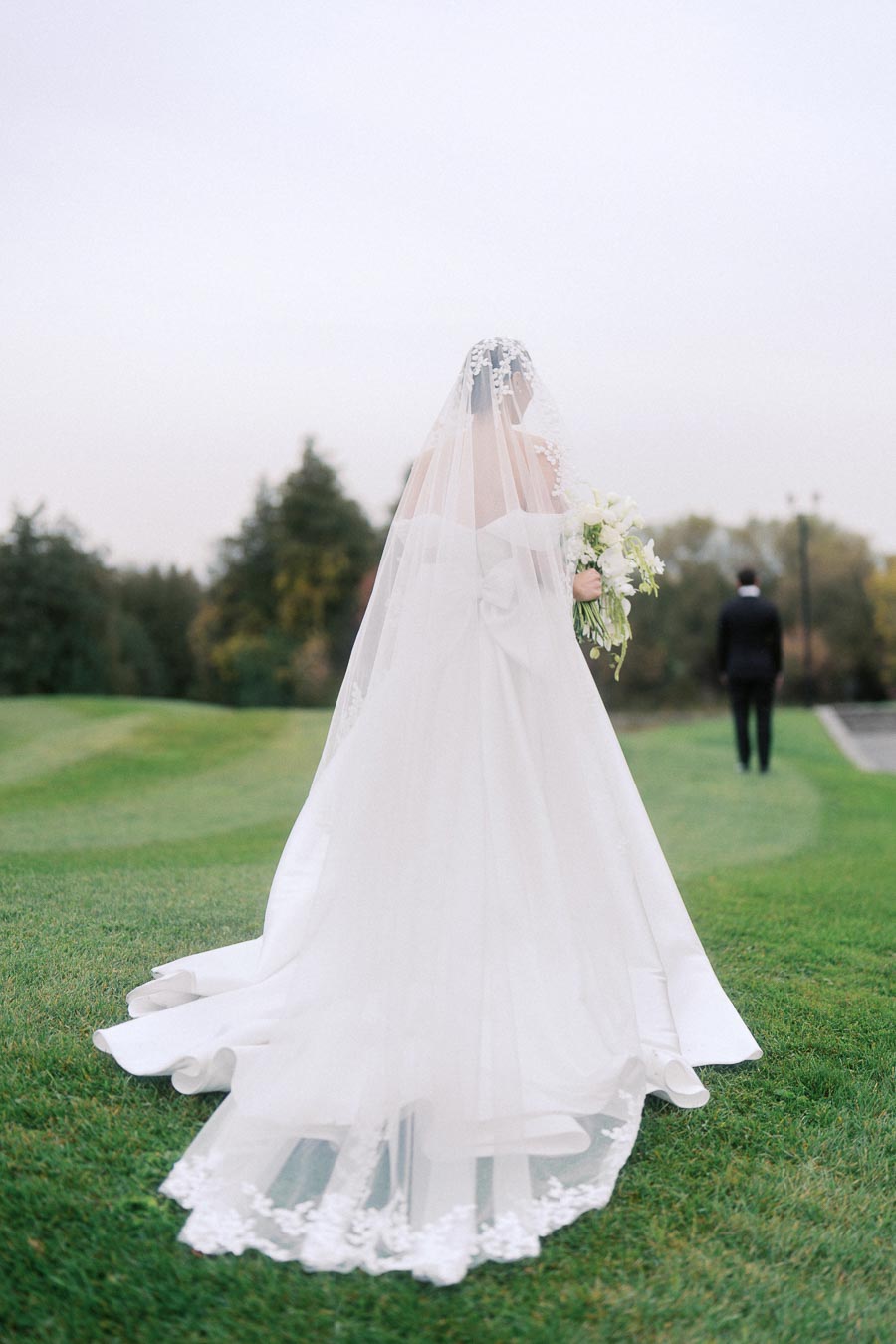 Elegant bride in a flowing white gown and veil holding a bouquet, walking towards the groom on a lush green lawn, perfect wedding day setting.
