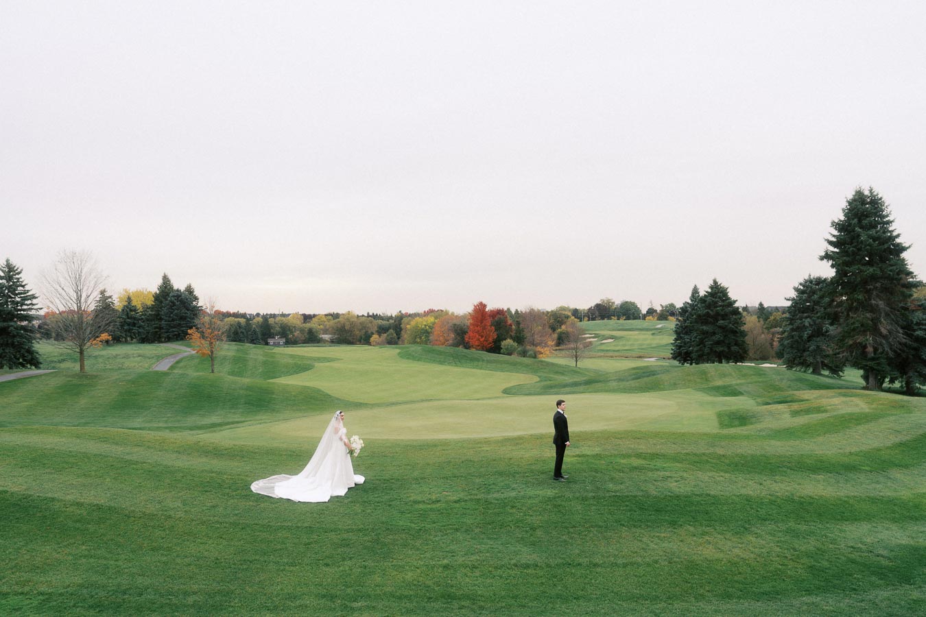 Bride in elegant gown walking towards groom on a golf course with lush green landscape and autumn trees.