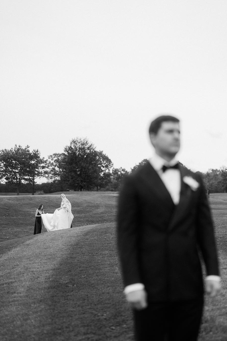 Black and white image of a groom in focus in the foreground, with a bride in a wedding dress and attendant adjusting her train in the background on a grassy field with trees.