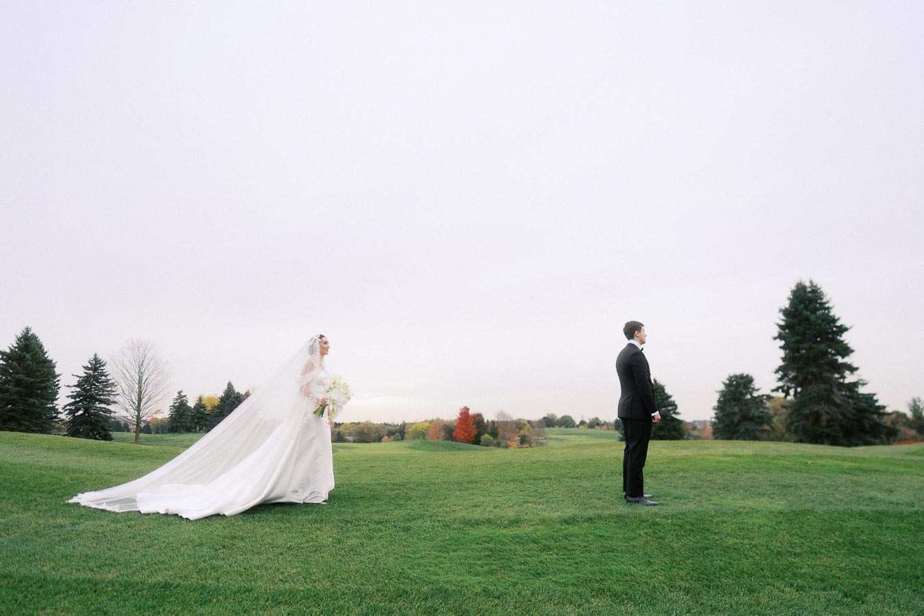 Bride in a long flowing gown approaching groom on a scenic golf course, surrounded by lush greenery and autumn trees.