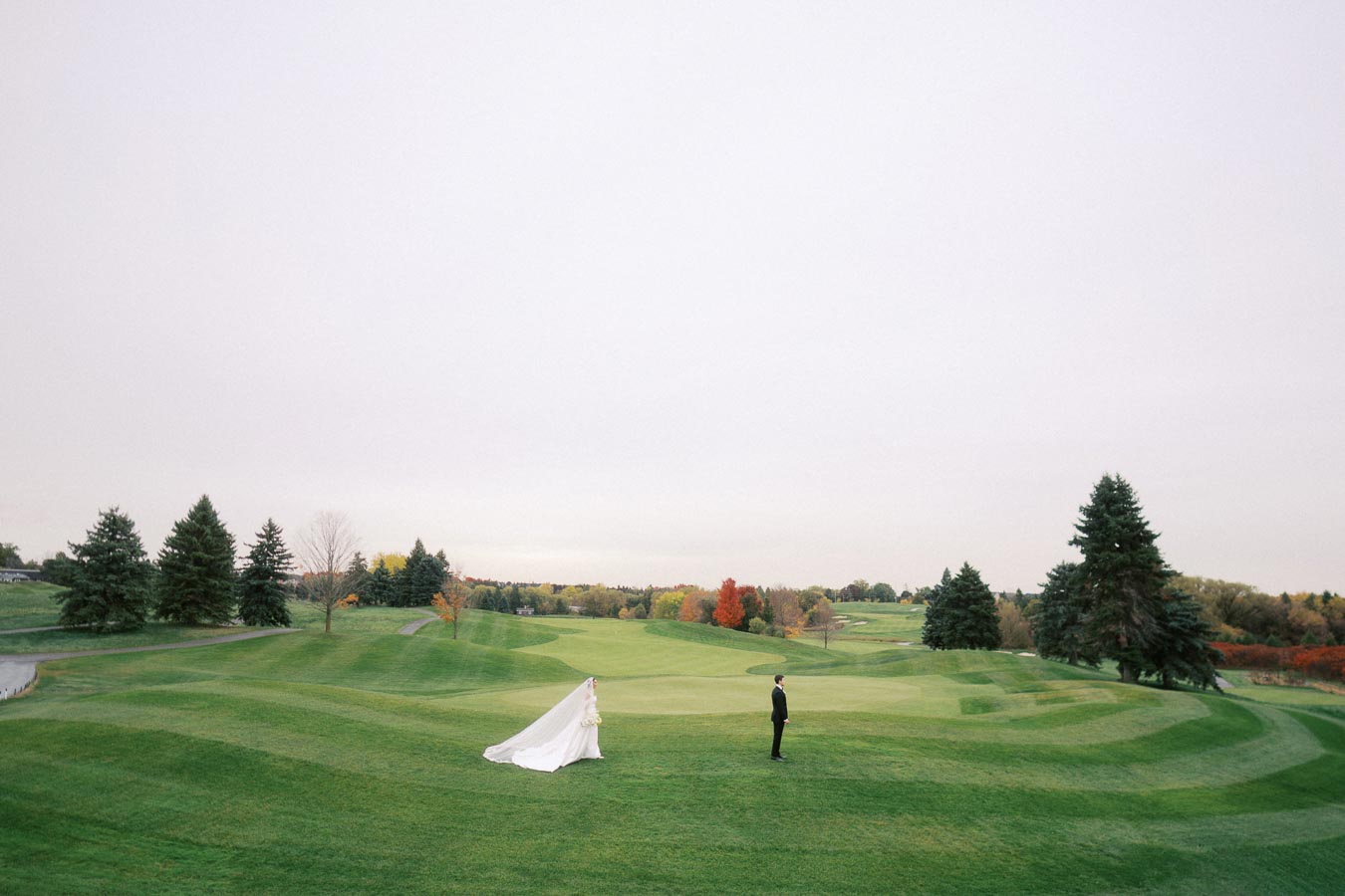 Bride and groom stand on a lush green golf course surrounded by autumn trees, with the bride's long veil flowing behind her.