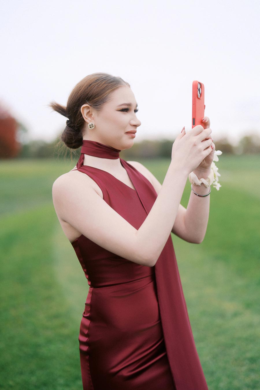 Young woman in a burgundy dress taking a photo with a smartphone in a scenic outdoor setting.