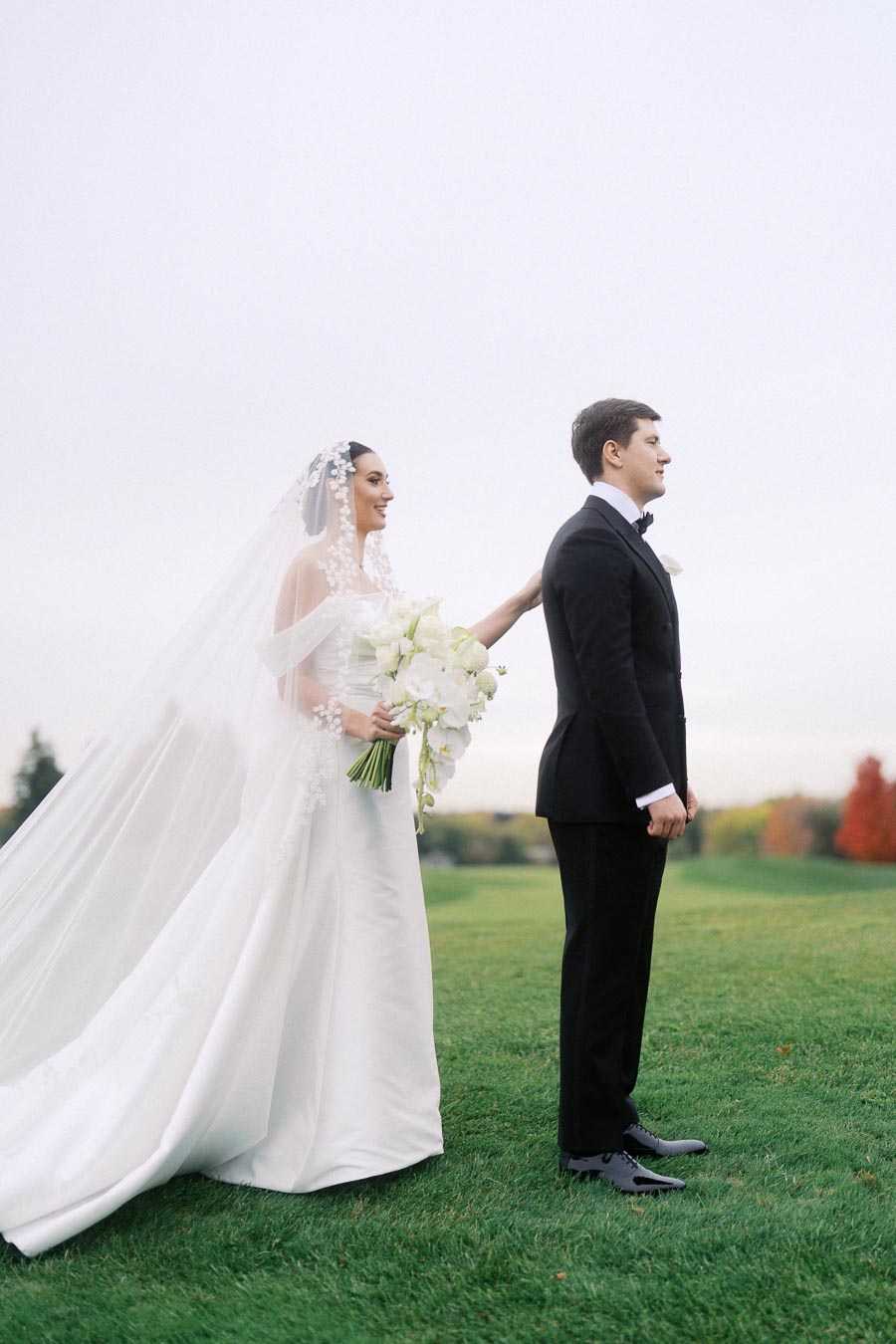 Bride in elegant white gown with floral veil, holding a bouquet, approaching groom in black tuxedo on a lush green lawn during wedding first look moment.