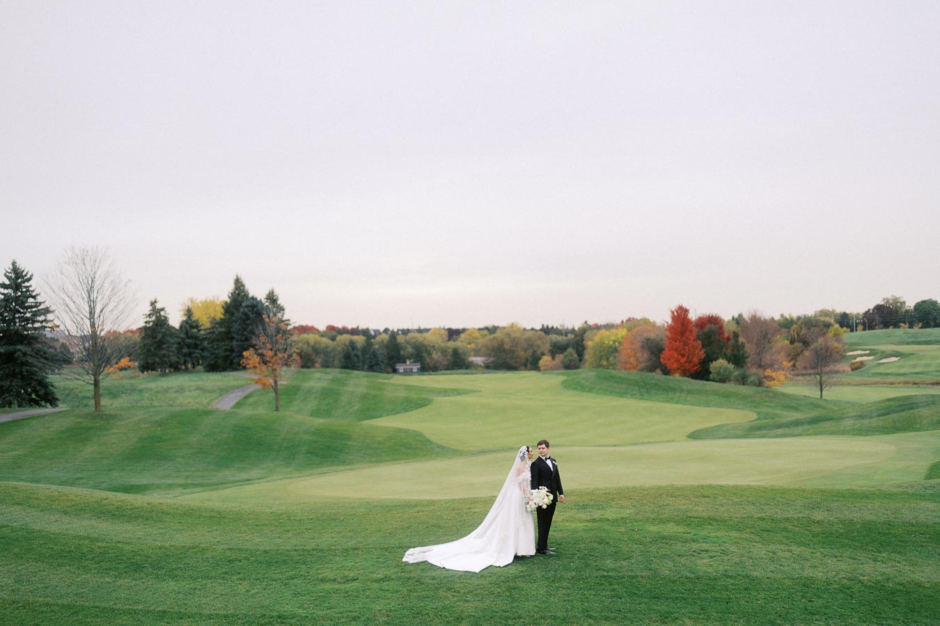 A bride and groom stand on a lush green golf course with vibrant autumn trees in the background, creating a picturesque wedding scene.