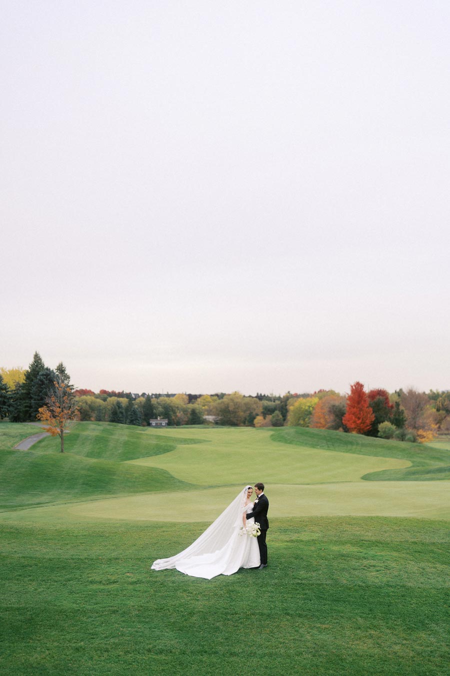 A bride and groom stand on a lush green golf course, surrounded by vibrant autumn trees, captured under an overcast sky.