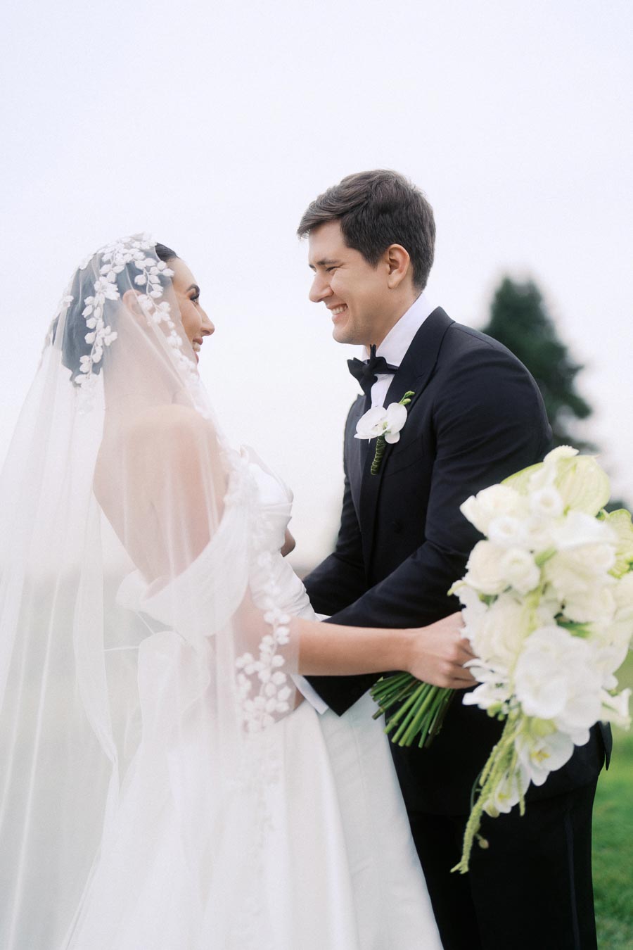 A bride and groom share a joyful moment outdoors; the bride is in a white gown with a veil, holding a bouquet of white flowers, while the groom wears a black tuxedo with a white boutonniere, against a blurred natural backdrop.