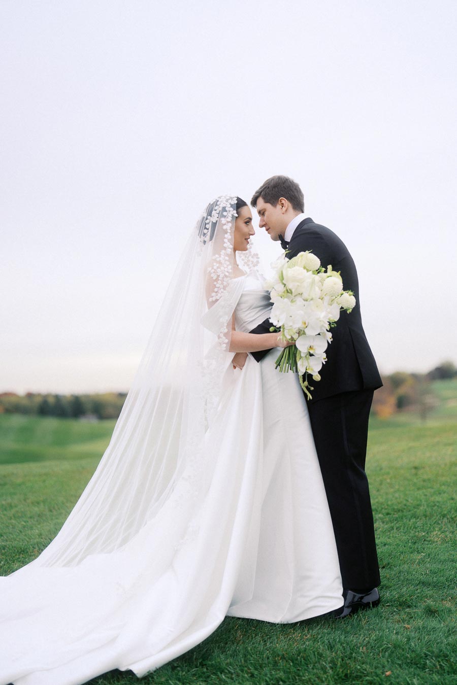 Bride and groom standing together on a grassy field, with the bride wearing a flowing white dress and veil, and the groom in a black suit, holding a bouquet of white flowers.
