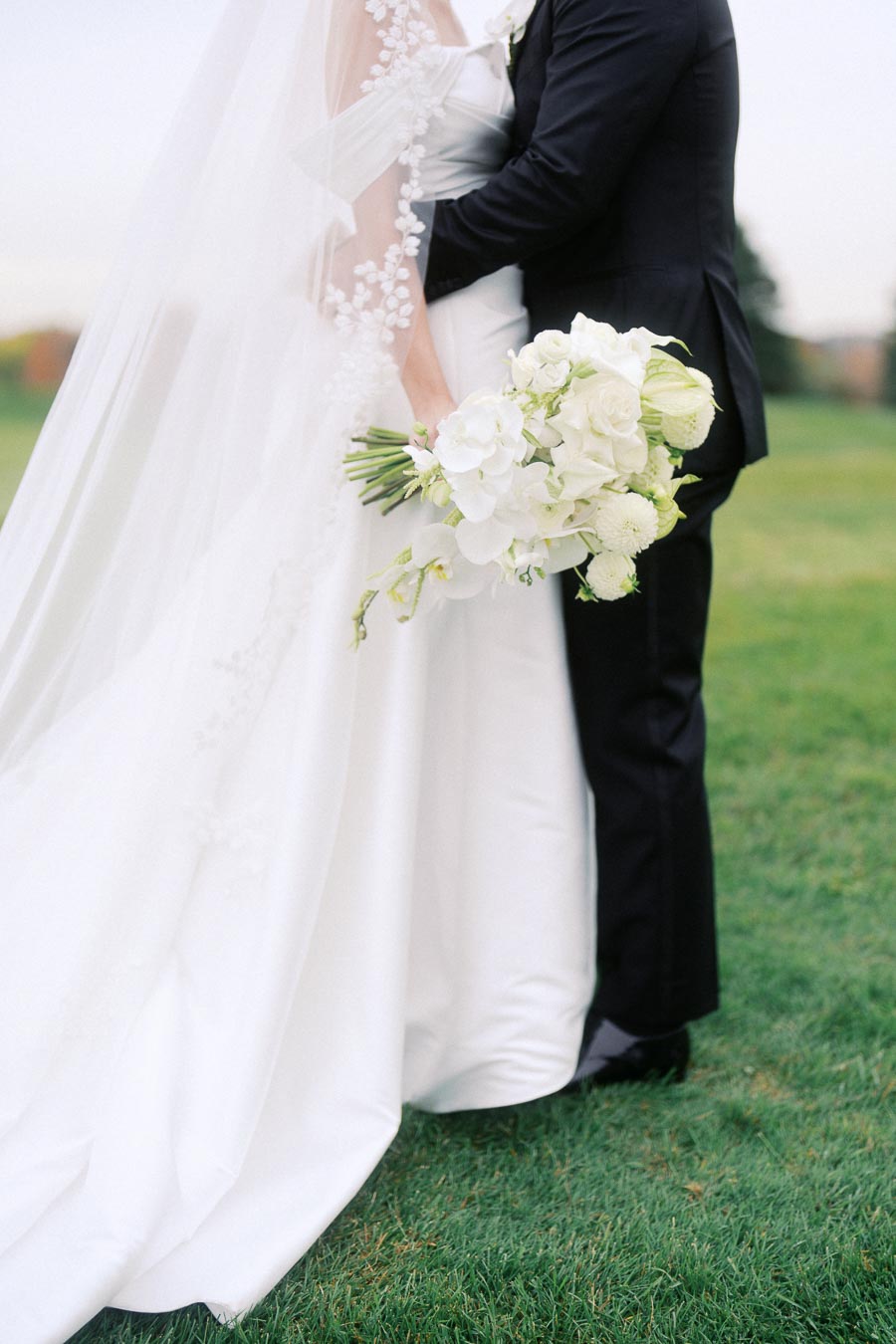 Elegant bride in a white wedding gown holds a bouquet of white flowers, standing closely with the groom in a black suit on a lush green lawn.