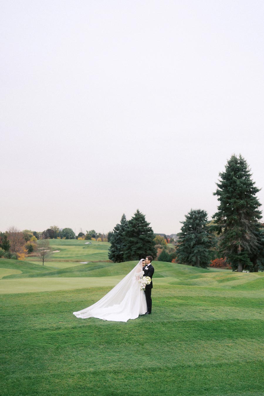 Bride and groom embrace on a scenic golf course, surrounded by lush greenery and tall trees, under an overcast sky.