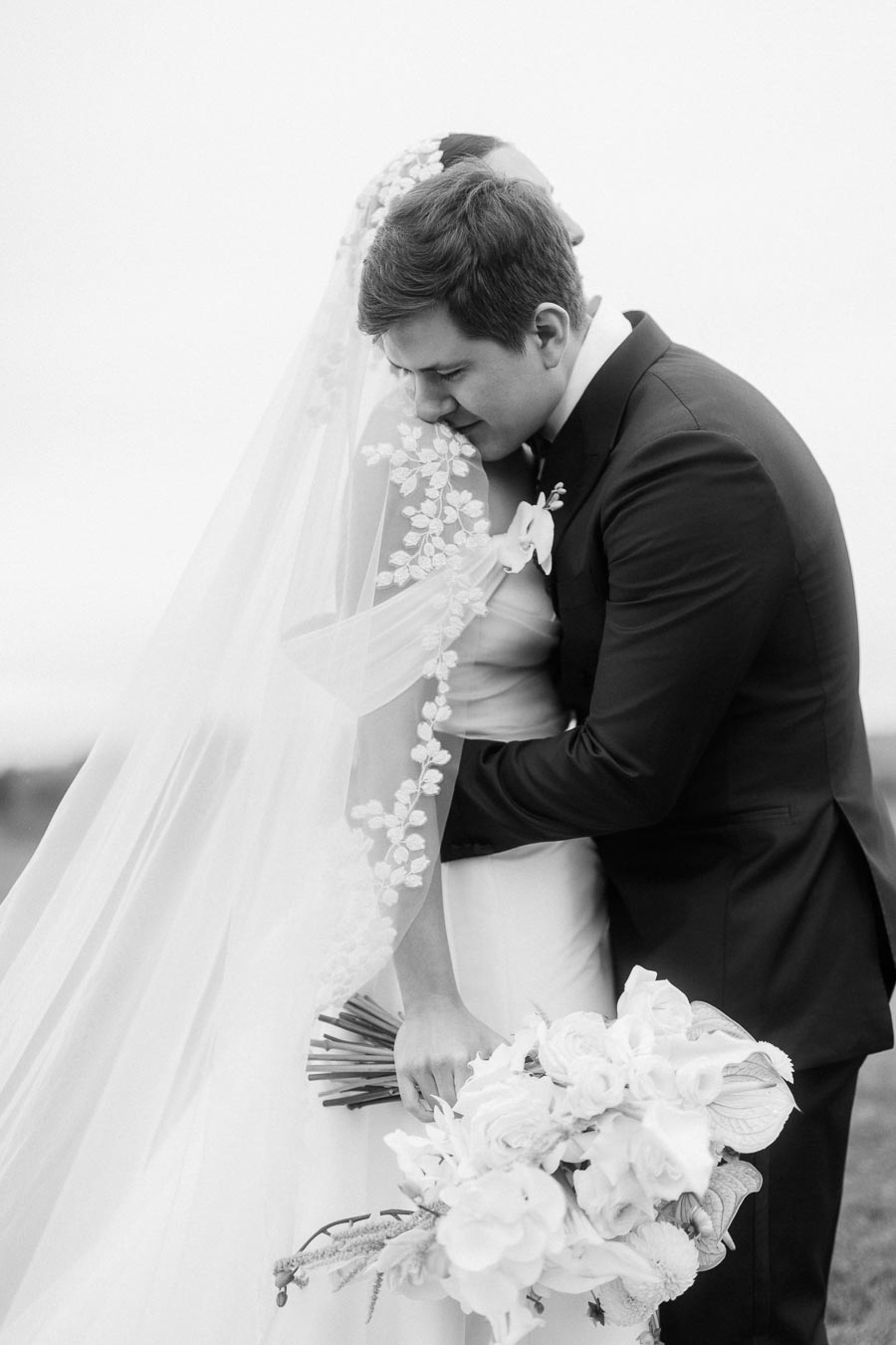 Black and white photo of an embracing bride and groom on their wedding day, with the bride holding a bouquet of white flowers and wearing a veil adorned with floral designs.