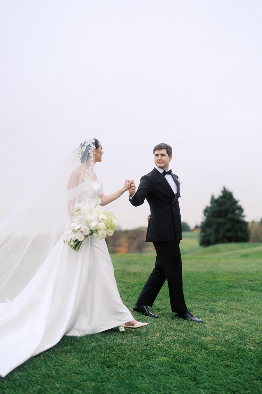 Bride and groom holding hands in a picturesque outdoor setting, the bride wearing a stunning white gown and veil with a bouquet, and the groom in a classic black tuxedo, walking together on lush green grass.