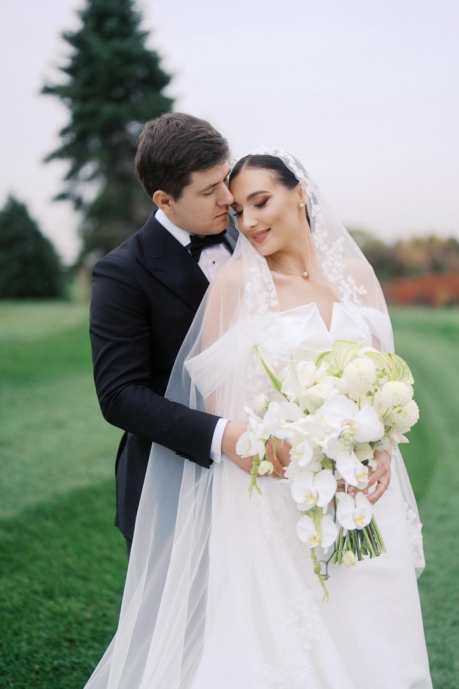 Bride and groom embracing on their wedding day, with the bride holding a bouquet of white flowers and the groom in a black suit, surrounded by a picturesque outdoor setting with green grass and trees.