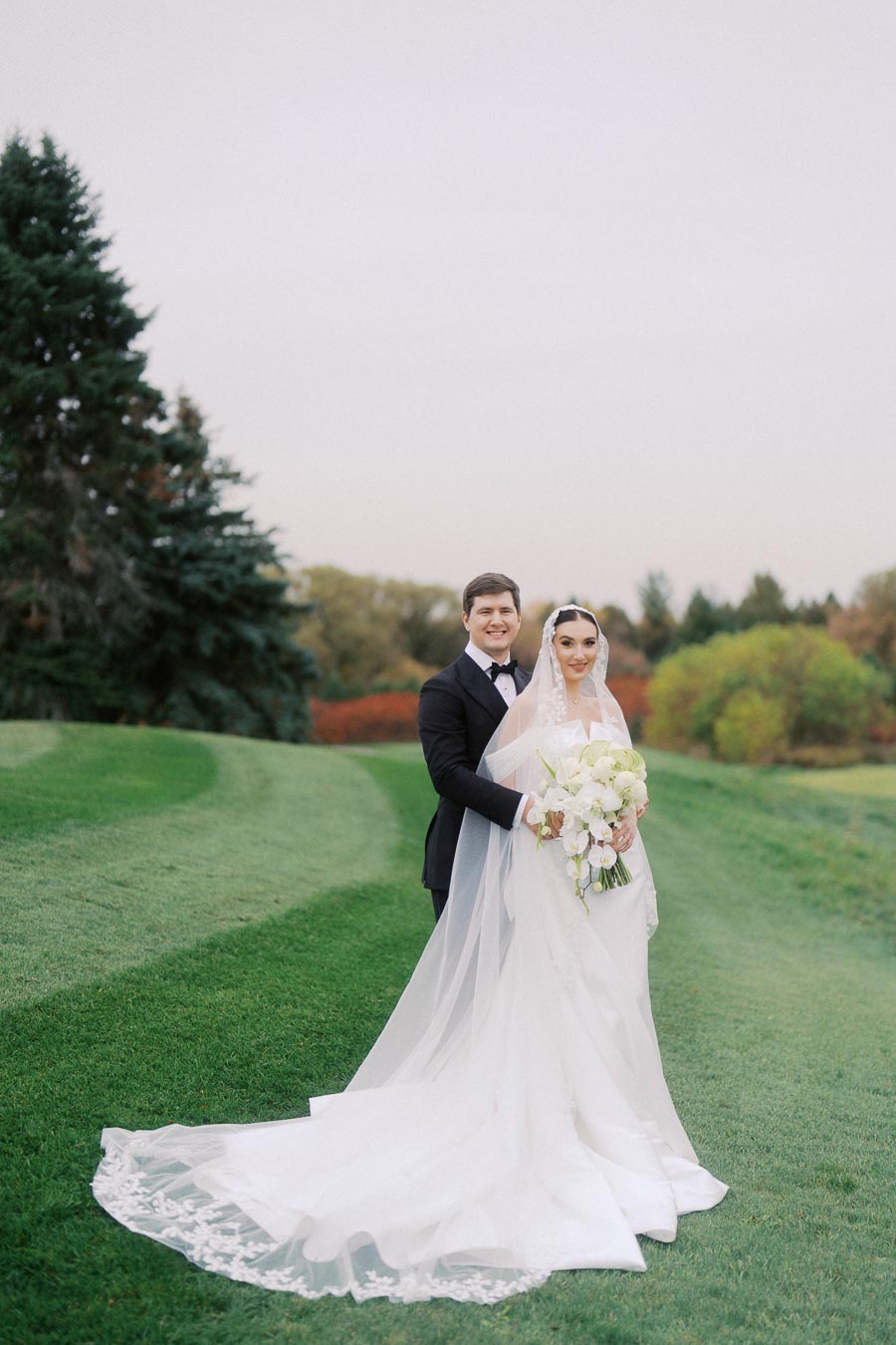 Bride and groom posing outdoors on a lush green lawn, with the bride in a long white dress holding a bouquet and the groom in a black tuxedo, against a backdrop of trees and a clear sky.