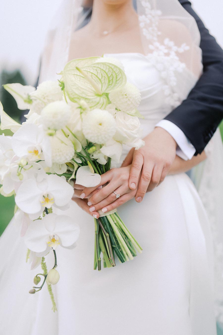 Bride holding a bouquet of white flowers with groom embracing her from behind, showcasing wedding ring and elegant bouquet.