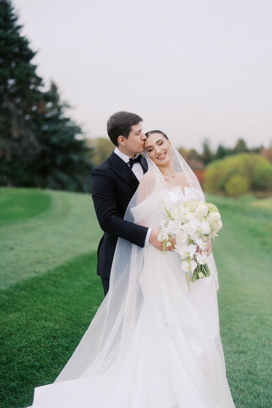 A bride and groom embrace on their wedding day, standing on a lush green lawn with a backdrop of trees. The groom, in a black tuxedo, kisses the bride, who is wearing a white gown and holding a bouquet of white flowers, capturing a moment of joy and romance.