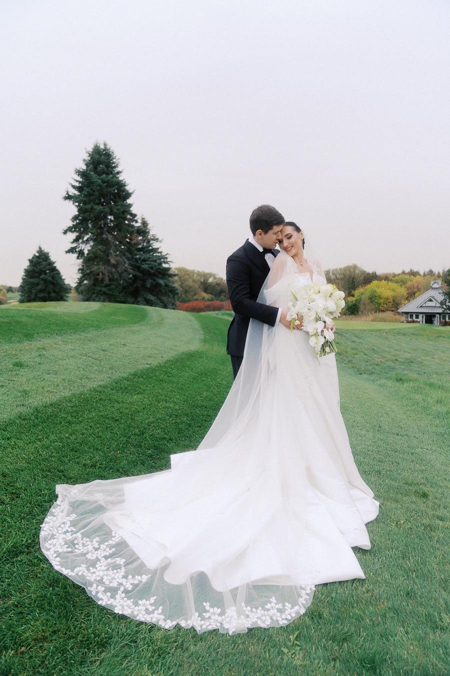 Elegant bride and groom embracing on a lush green lawn with a flowing white wedding gown, surrounded by vibrant autumn trees, capturing a perfect outdoor wedding moment.