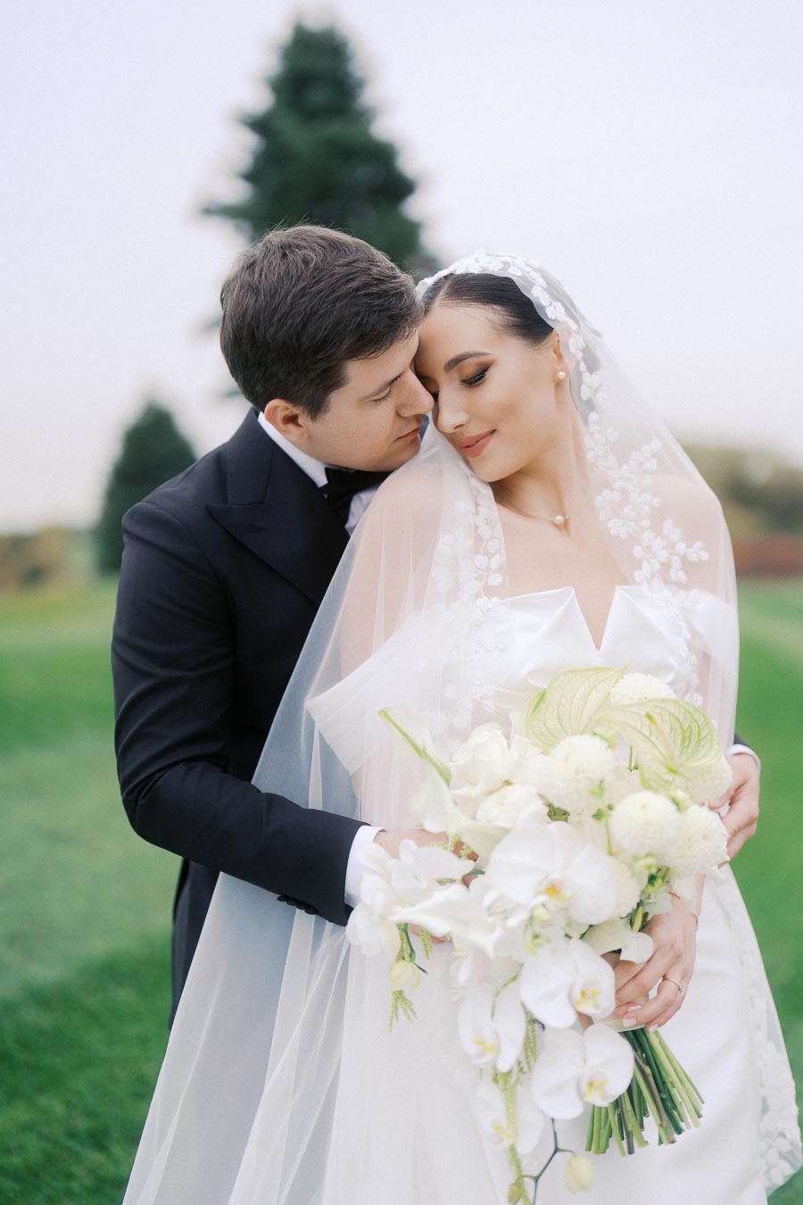 Bride and groom embracing outdoors on their wedding day, with the bride holding a bouquet of white flowers and wearing a floral veil.