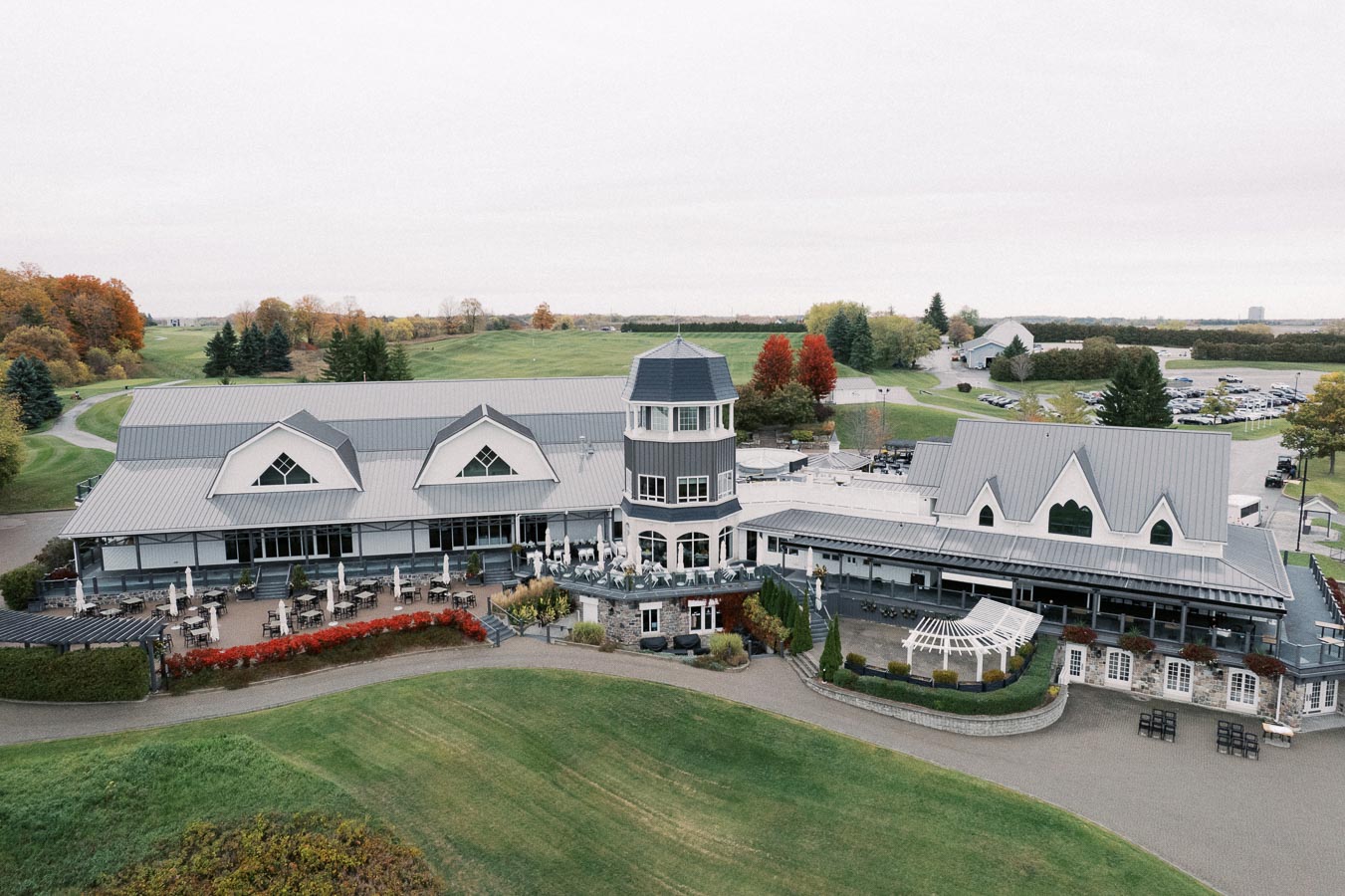 Aerial view of a large, elegant clubhouse surrounded by lush green golf course landscape, featuring a distinctive tower and spacious outdoor seating area, with autumn trees in the background.