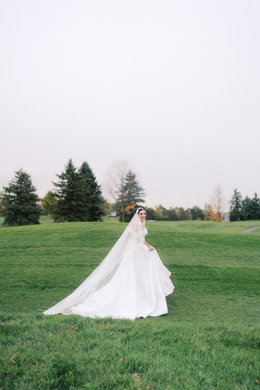 Bride in a flowing white wedding gown and veil stands on lush green grass with a serene backdrop of trees and a cloudy sky.