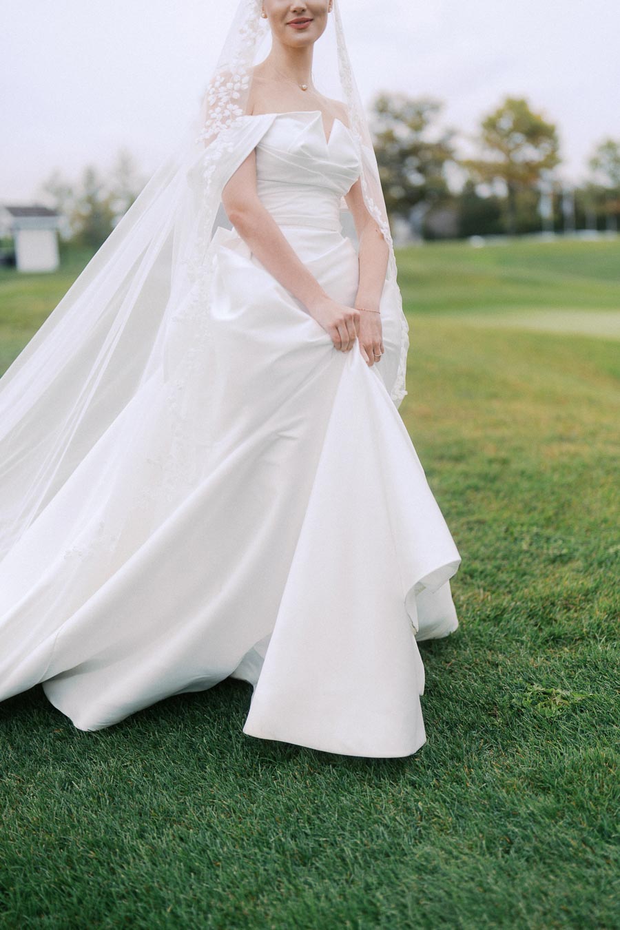 Bride in an elegant white wedding gown with lace details and a flowing train, standing on lush green grass outside, showcasing a classic and romantic bridal look.