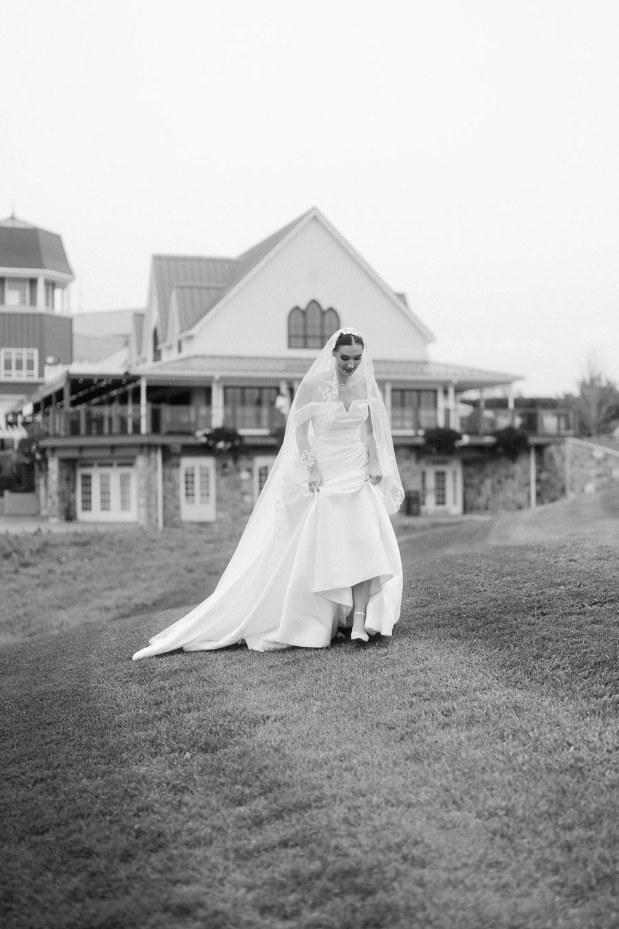 Bride in elegant white wedding gown and veil walking on grassy lawn in front of a charming building, showcasing a timeless and romantic wedding scene.