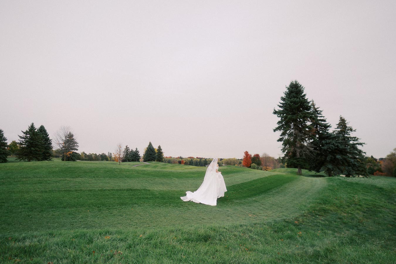 Bride walking gracefully on a lush green field surrounded by tall trees, creating a serene and scenic wedding setting.