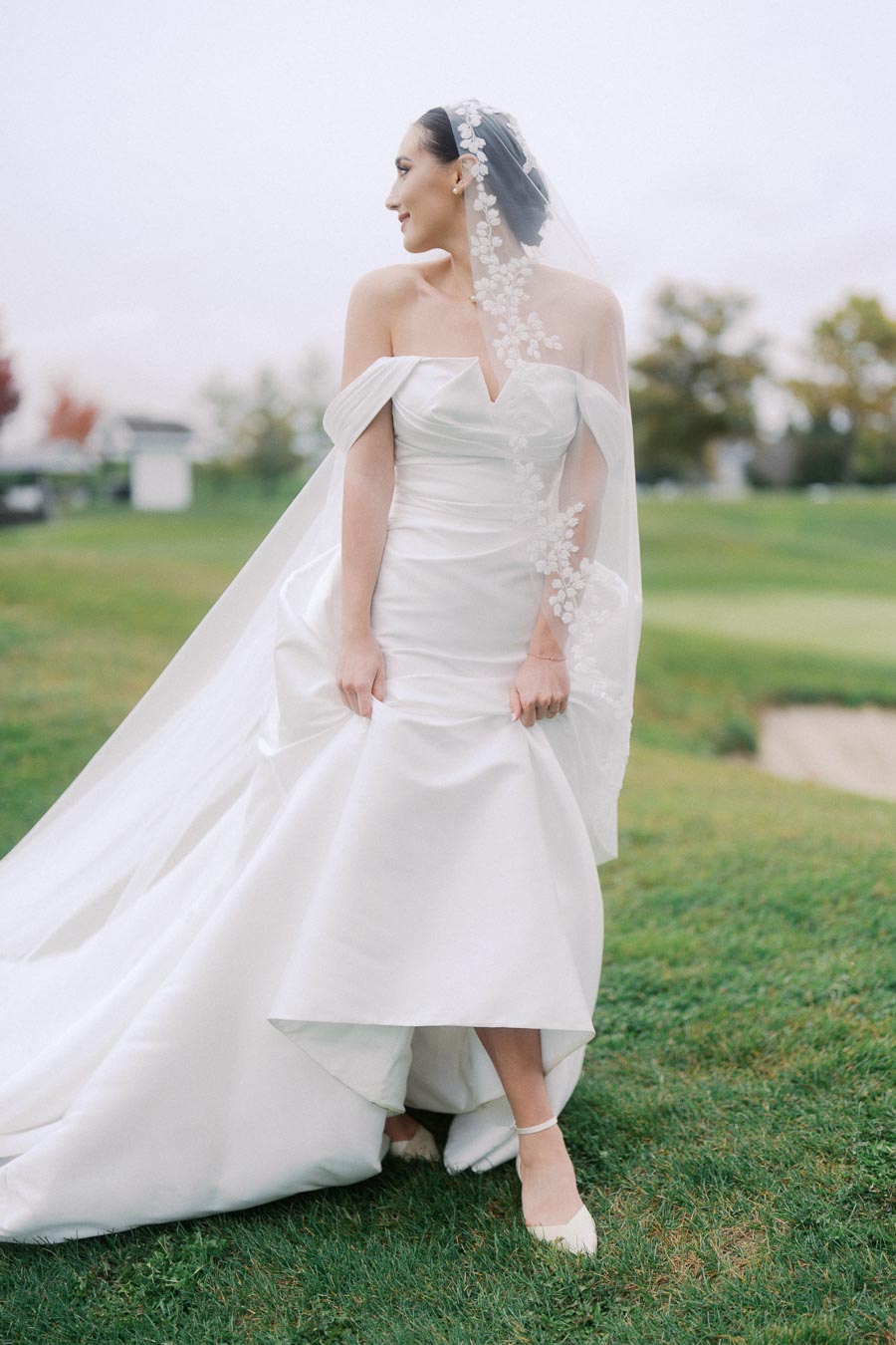 Bride in elegant white wedding gown with lace veil and off-the-shoulder design, standing on a grassy field.