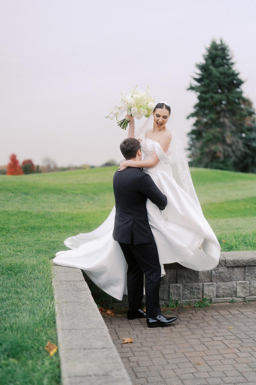 Bride and groom celebrating outdoors, the bride in a flowing white gown holding a bouquet, and the groom lifting her joyfully, surrounded by lush greenery and a brick pathway.