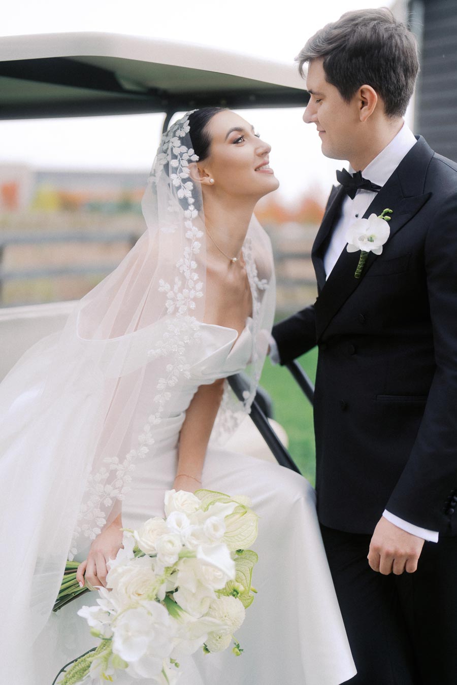 Bride in elegant white wedding gown and veil shares a joyful moment with groom in sharp black tuxedo, holding a bouquet of white flowers, outside on a grassy area.