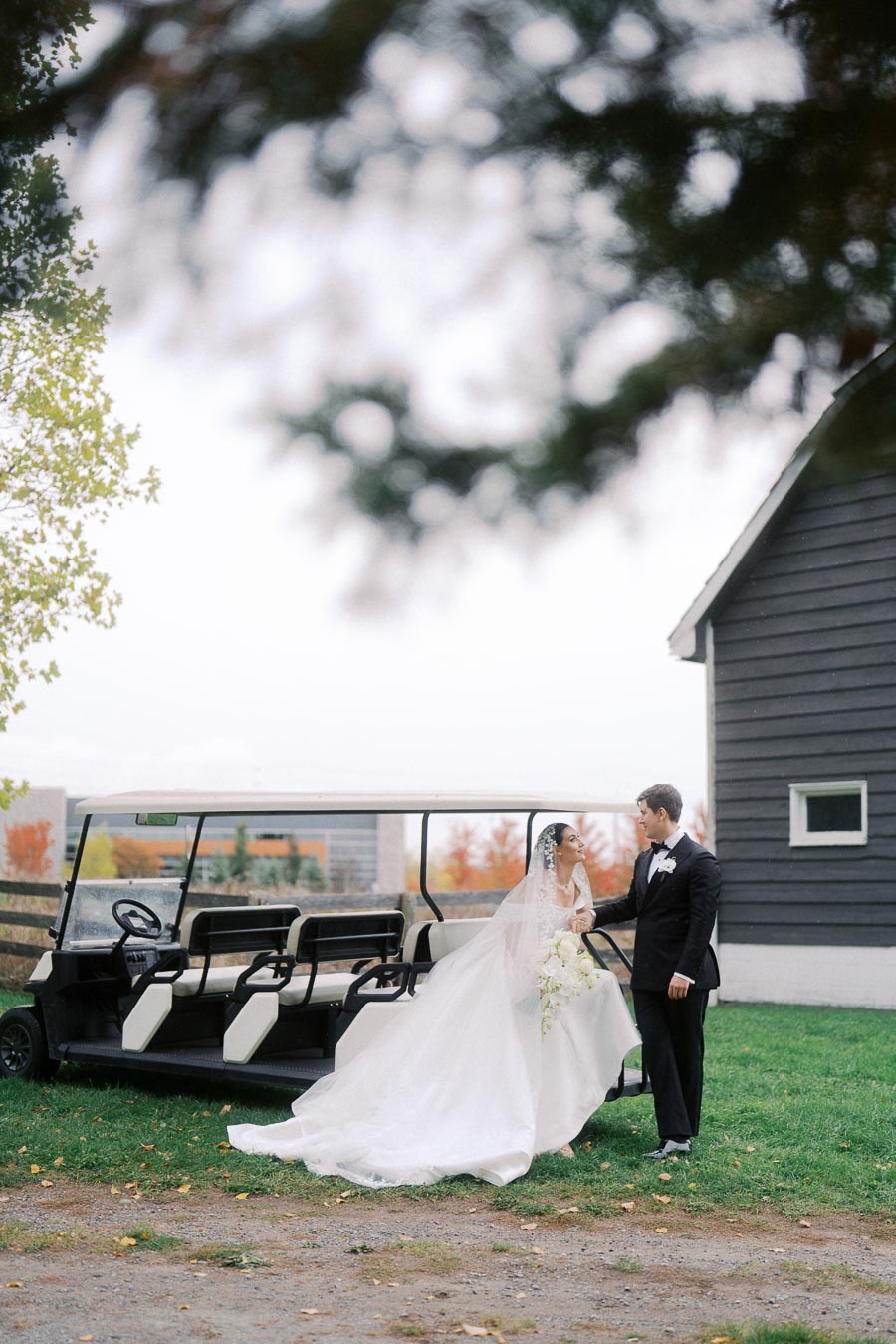 Bride and groom standing beside a golf cart in a scenic outdoor setting, with the bride in a flowing white dress holding a bouquet of white flowers, and the groom in a classic black tuxedo, near a rustic barn with autumn foliage in the background.