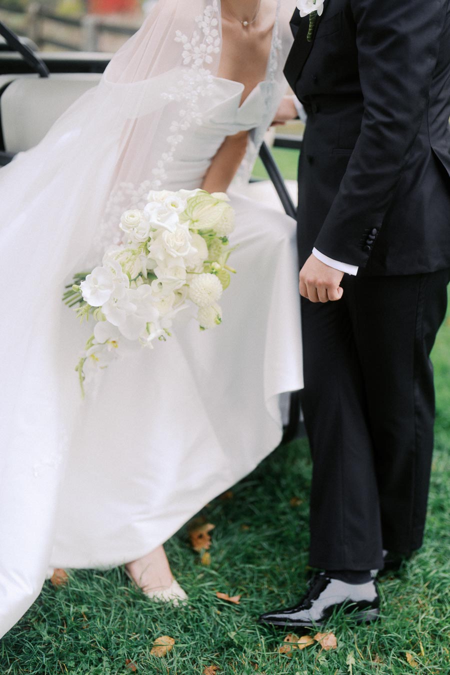 Bride and groom in elegant attire standing on grass, bride holding a bouquet of white flowers with greenery, highlighting wedding elegance and celebration.