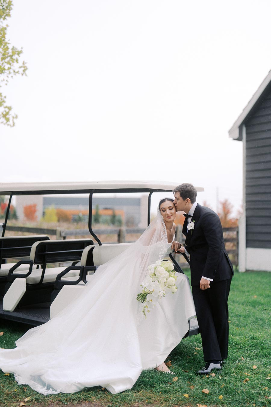 A bride and groom share a tender moment next to a golf cart on their wedding day, with the bride wearing a flowing white gown and holding a bouquet of white flowers. The groom is in a black suit, gently kissing her forehead. The background features autumn foliage and a rustic barn setting.