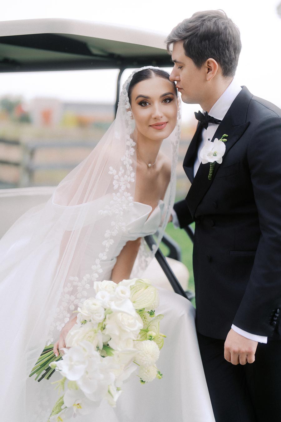 Bride in elegant white gown and veil holding a bouquet of white flowers, standing next to groom in black tuxedo with boutonniere, sharing an affectionate moment by a vehicle on their wedding day outdoors.