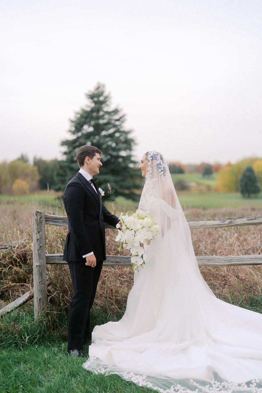 Bride and groom standing in a scenic outdoor setting with a wooden fence, bride in a white gown and veil holding an elegant bouquet of white flowers, and groom in a black tuxedo, lush green field and trees in the background.