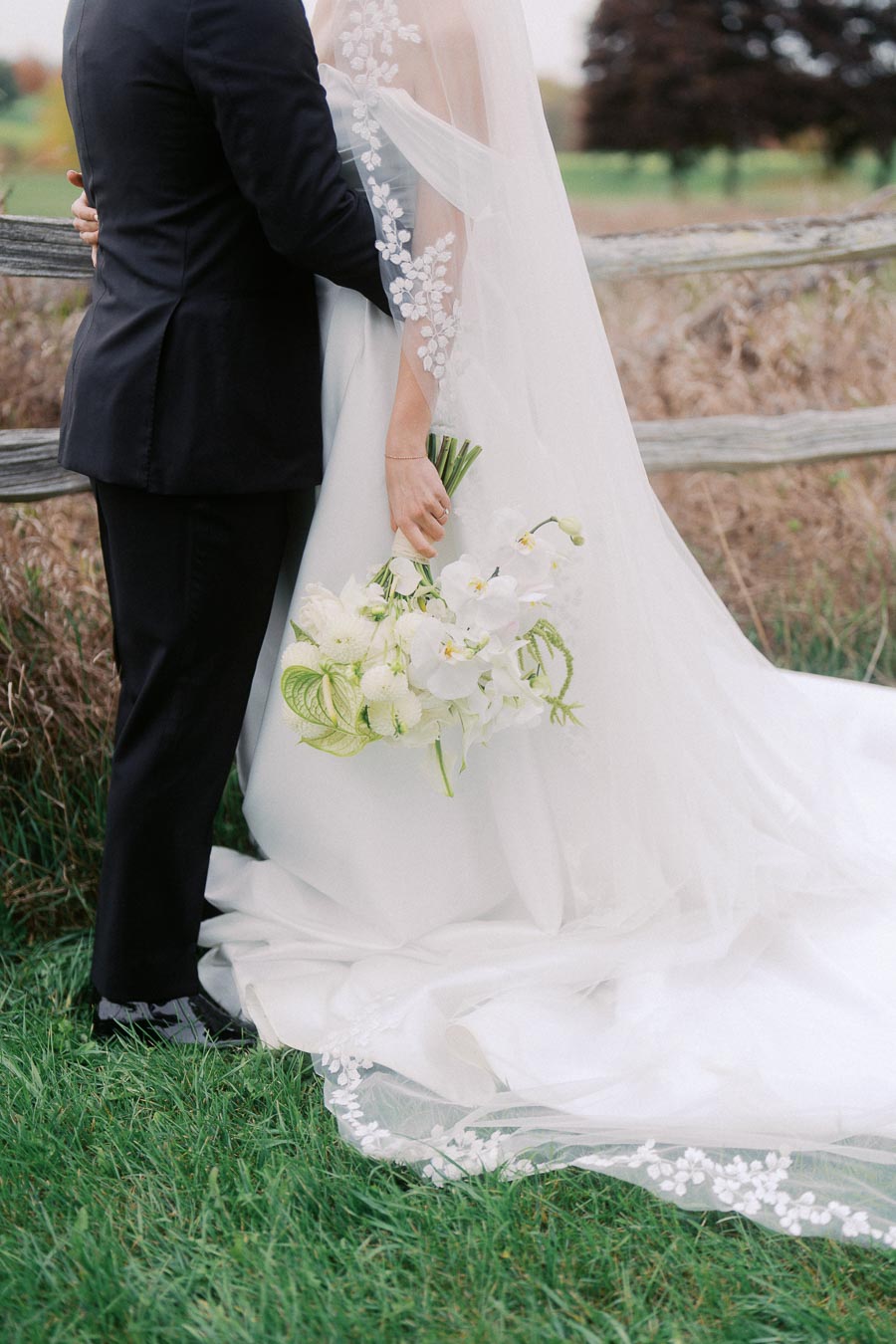 Bride and groom embracing near rustic wooden fence, with bride holding elegant bouquet of white flowers and wearing a delicate floral-embroidered veil, in a picturesque outdoor wedding setting.